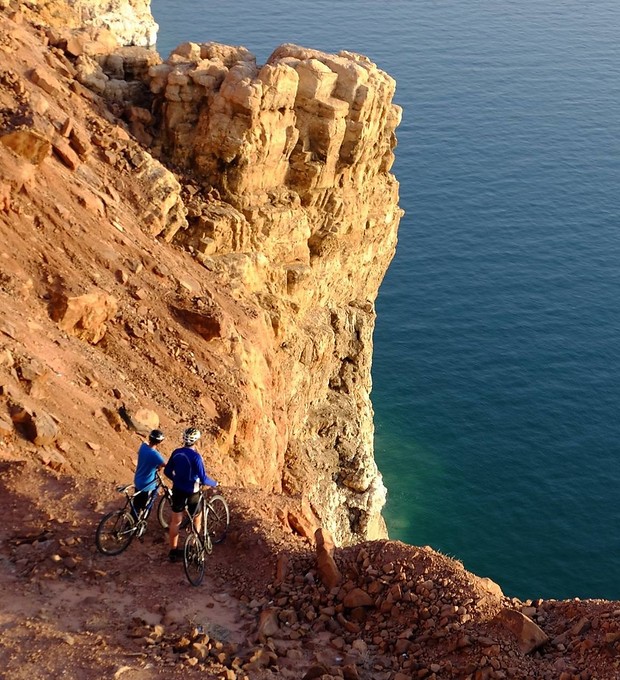 Two cyclists pause on a narrow, rocky cliff edge overlooking a vast, calm ocean, surrounded by rugged, reddish terrain. They wear helmets and observe the scene below.